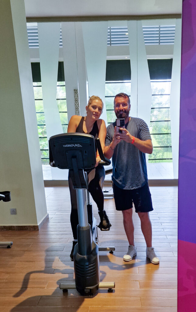 Luke and Kay at the riu hotel sri lanka gym with Kay on the exercise bike and Luke standing up taking pic on phone