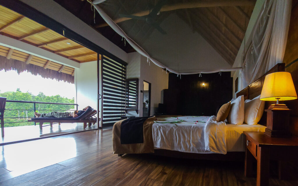 A large double bed in the premium deluxe room at 98 Acres Resort and Spa Hotel and Luke sitting on a sun lounger on a balcony looking over the tea plantations