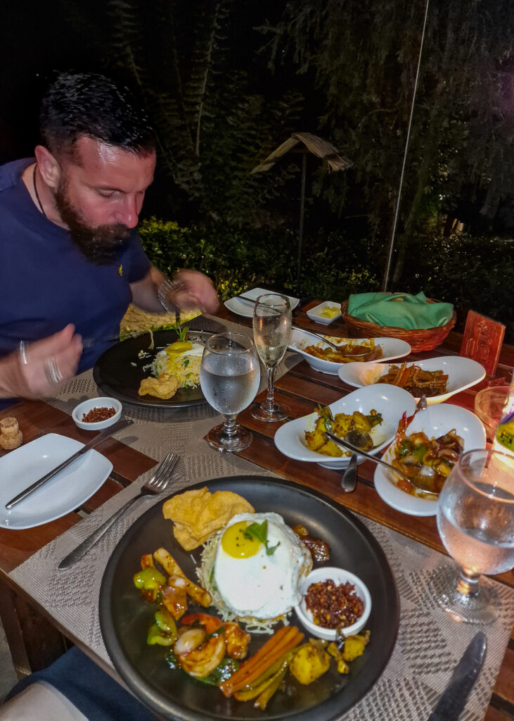 Luke eating his dinner at the restaurant 98 at 98 acres resort in Ella Sri Lanka. 98 acres oriental set meal.  Stir-fried noodles topped with a fried egg. To the side, there's a spicy serving of devilled prawns and calamari. Accompaniments include a tempered potato dish, a vegetable chop suey, and crispy prawn crackers. The meal is completed with a fiery chilli paste and a sweet mango chutney 