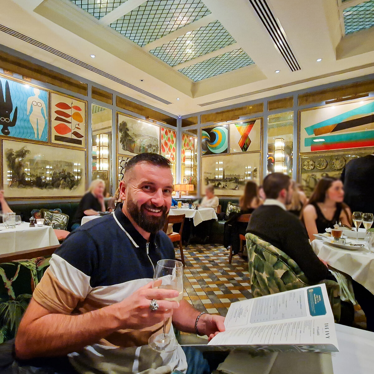 Luke from Flawless Journeys sitting in the main seating area holding a glass of wine and the menu at the Ivy brassiere in Bath